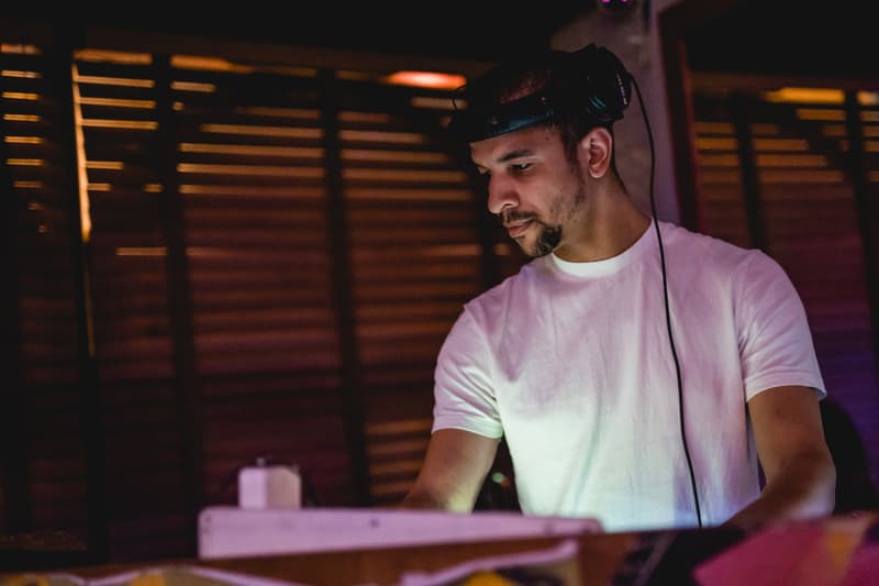 Austin Ventour in the DJ booth at night, headphones on, warm light through slatted blinds behind him
